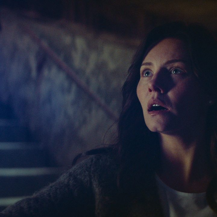 Keira looks tense as she stands at the bottom of the stairs in a scene from The Cellar