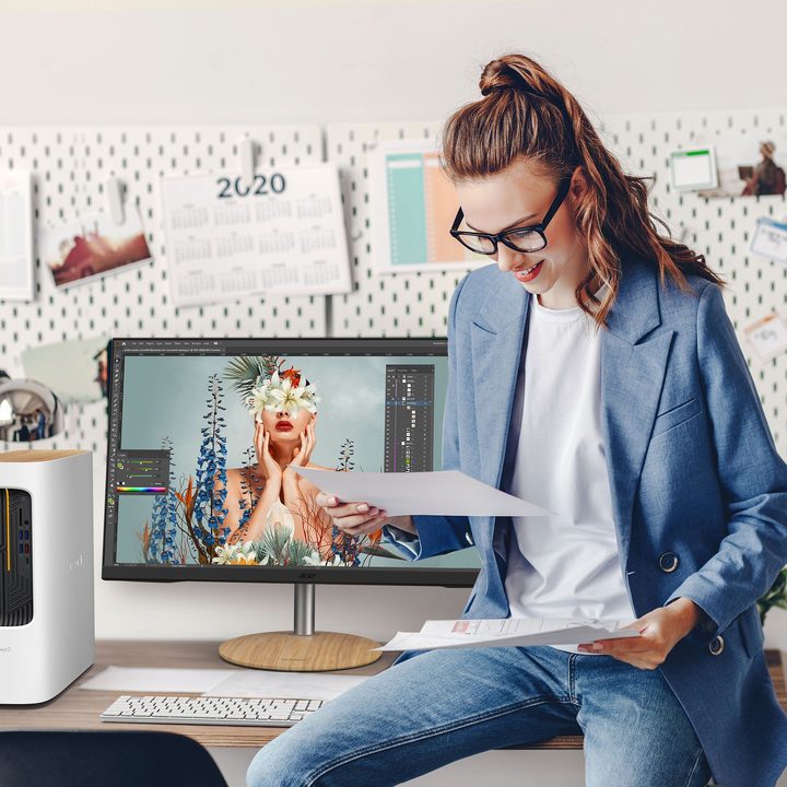A woman sits on a desk. Behind her, the Acer ConceptD workstation stands alongside a monitor.