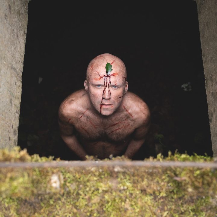 Rory Kinnear stares upward, naked, from a pit, with scratches on his body and a leaf on his head.
