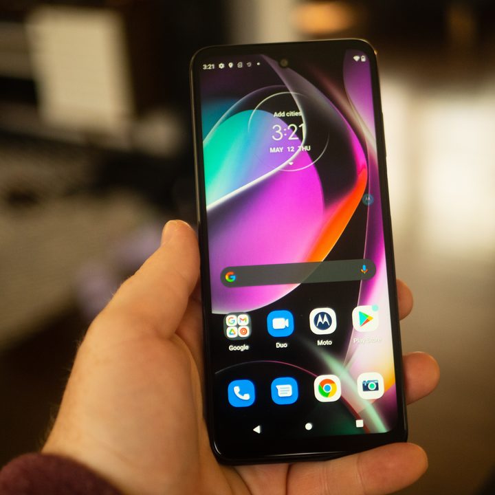 A hand holding the Moto G 5G smartphone with the homescreen displayed