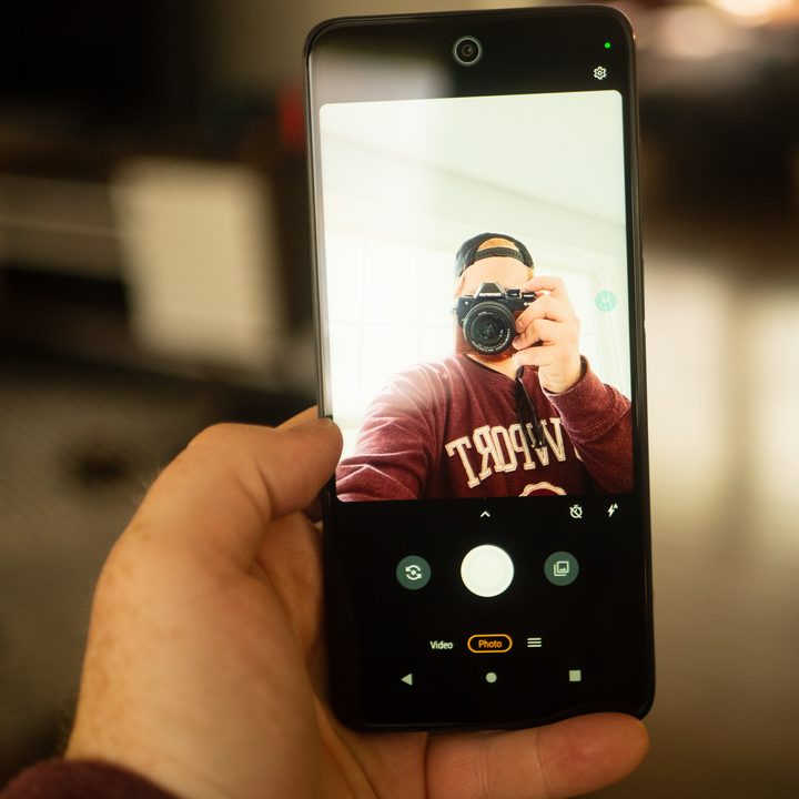 A hand holding the Moto G 5G smartphone with the selfie camera opened, depicting a man holding a separate camera