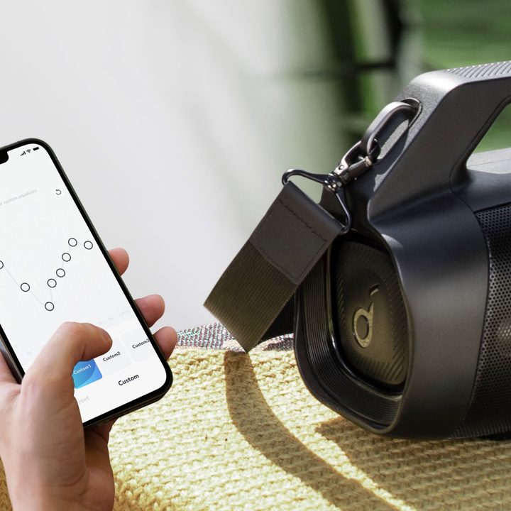 Soundcore Motion Boom Plus seen next to a hand holding a smartphone with the Soundcore app.