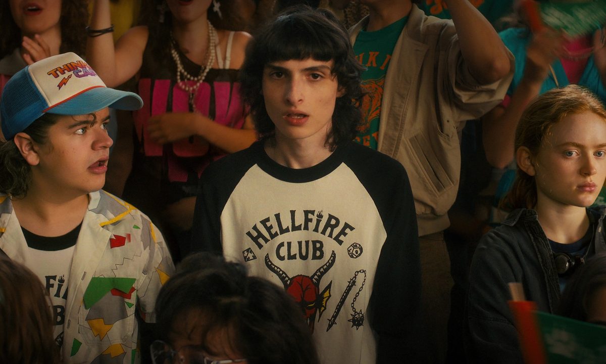 Gaten Matarazzo, Finn Wolfhard, and Sadie Sink stand in a crowd during a high-school rally in a scene from Stranger Things 4.