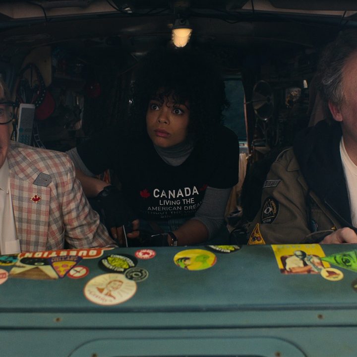 Two characters played by Mike Myers sit in the front seats of a van, with Lydia West peering out from behind them.