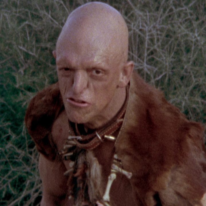 The mutant villain from The Hills Have Eyes is making a face in front of some brush.