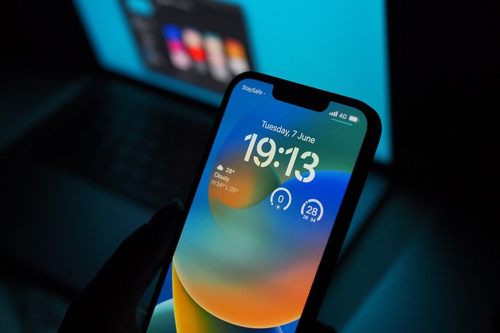 An iPhone 13 Pro running iOS 16 is held up in front of a Macbook.
