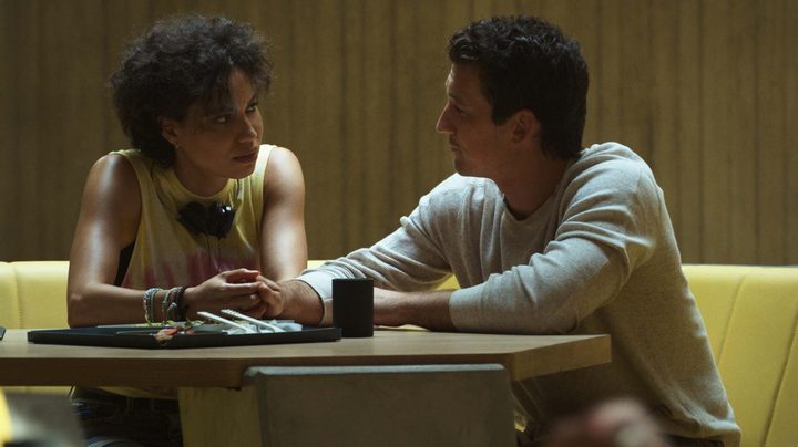 Jurnee Smollett and Miles Teller sit at a table, holding hands, in a scene from Spiderhead.