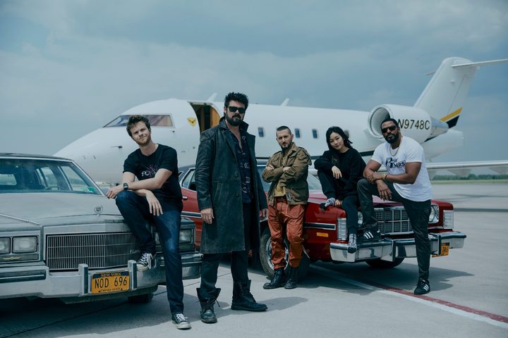 The cast of The Boys, with Karl Urban front and center, stand on an airstrip in front of a plane.