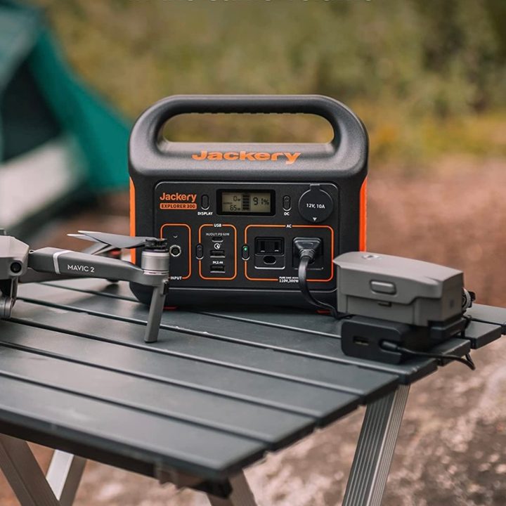 Jackery Portable Power Station Explorer 300 sits on a table at a camping site next to a drone.