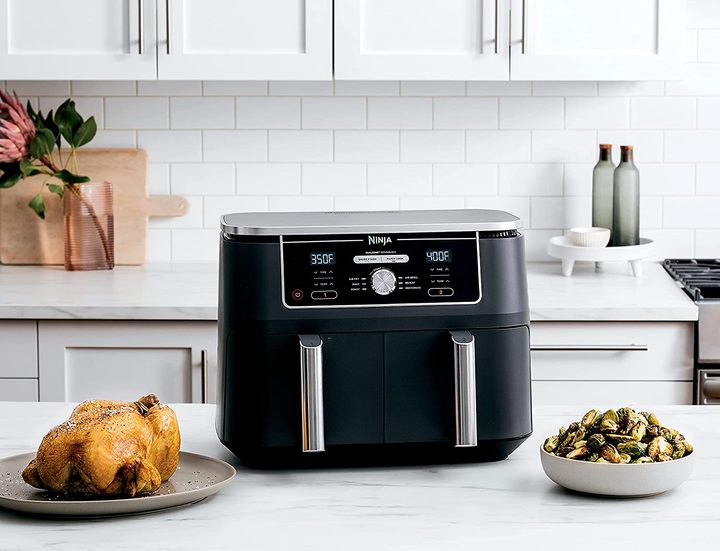 Ninja DZ401 Foodi 10 Quart 6-in-1 DualZone XL 2-Basket Air Fryer on a white kitchen counter with two cooked dishes.