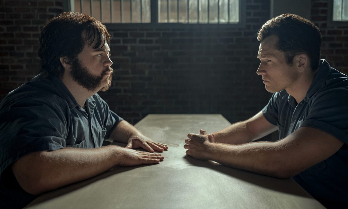 Paul Walter Hauser and Taron Egerton sit at a small table in prison, facing each other, in a scene from Black Bird.