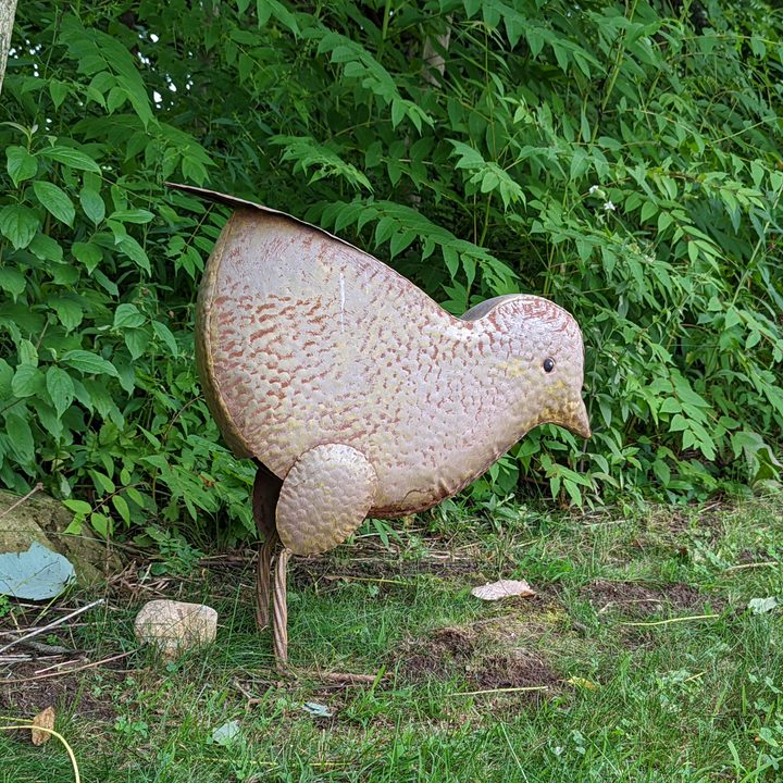 A large chicken decoration made out of metal. It's standing outside with a lot of greenery behind it.