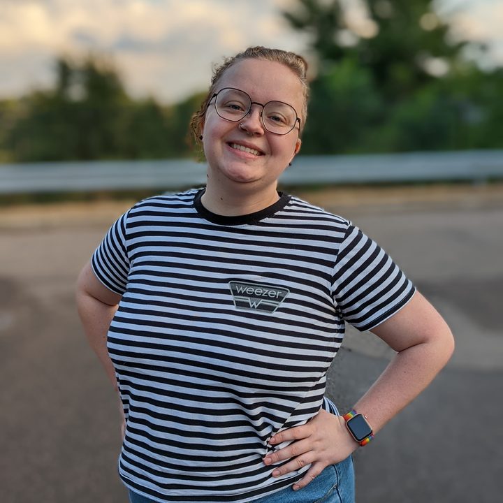 A person with blond hand, tied up in a bun. They're wearing a back and white striped shirt, hands on hips, and standing outside with a smile on their face.