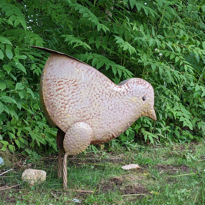 A large chicken decoration made out of metal. It's standing outside with a lot of greenery behind it.