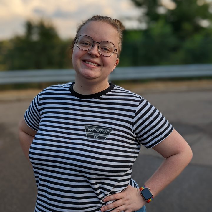 A person with blond hand, tied up in a bun. They're wearing a back and white striped shirt, hands on hips, and standing outside with a smile on their face.