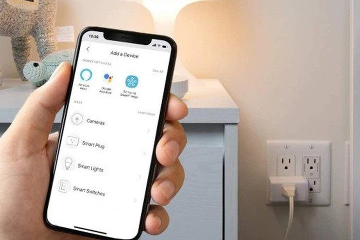 Smartphone in hand shows the Kasa Smart Plug app, with the plug powering a table lamp in the background.
