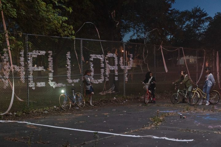 The four stars of Paper Girls stand next to a fence with "Hell Day" written on it.