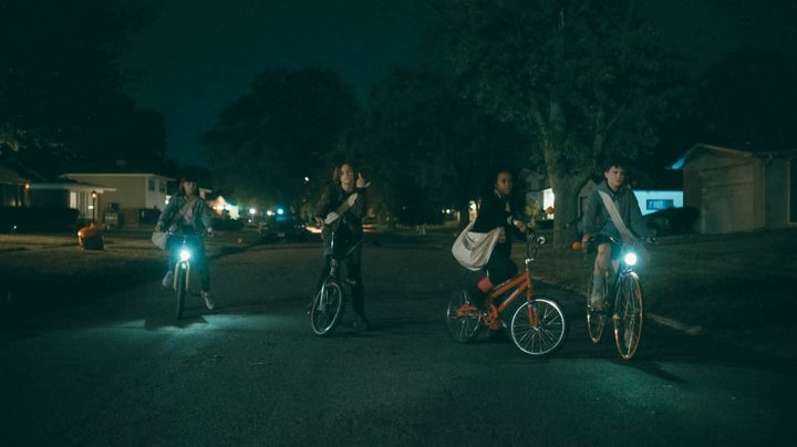 The four stars of Paper Girls sit on their bicycles on a dark street.