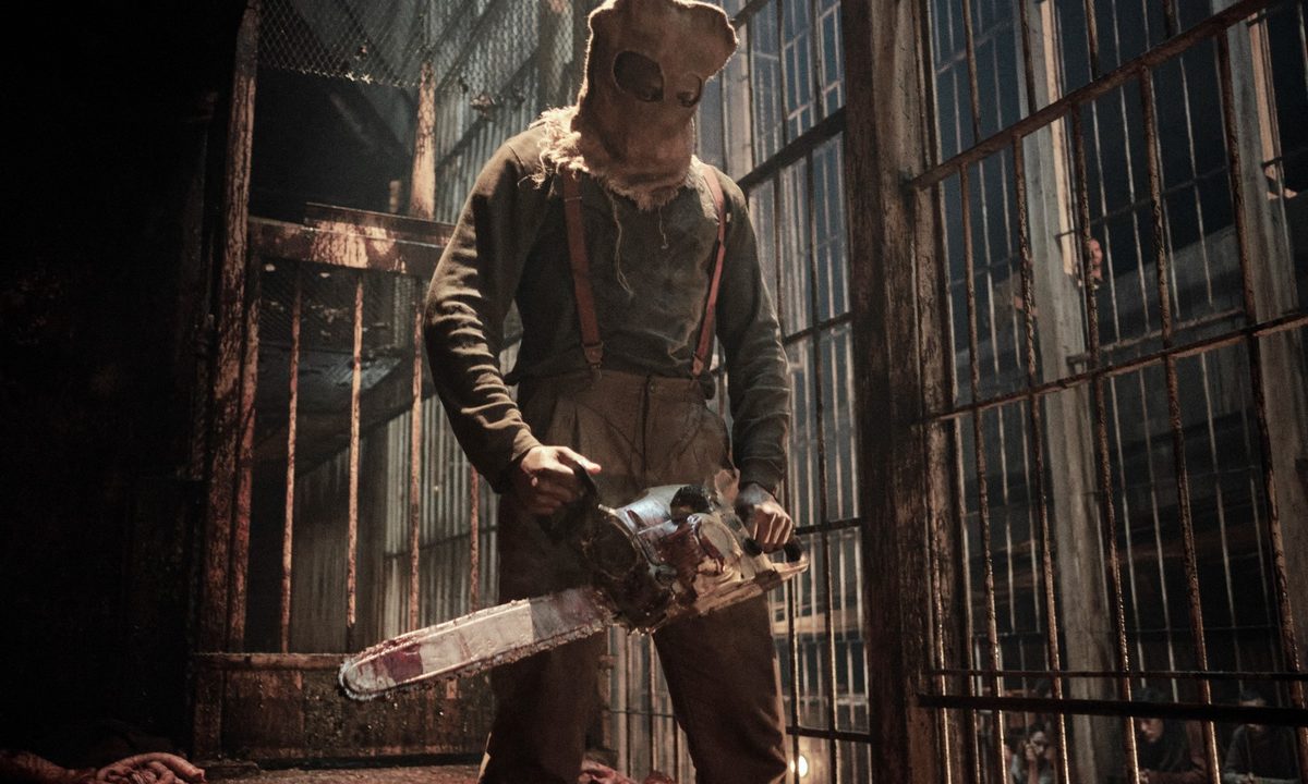A hooded figure holding a bloody chainsaw stands amid prison cells in a scene from the Resident Evil series.
