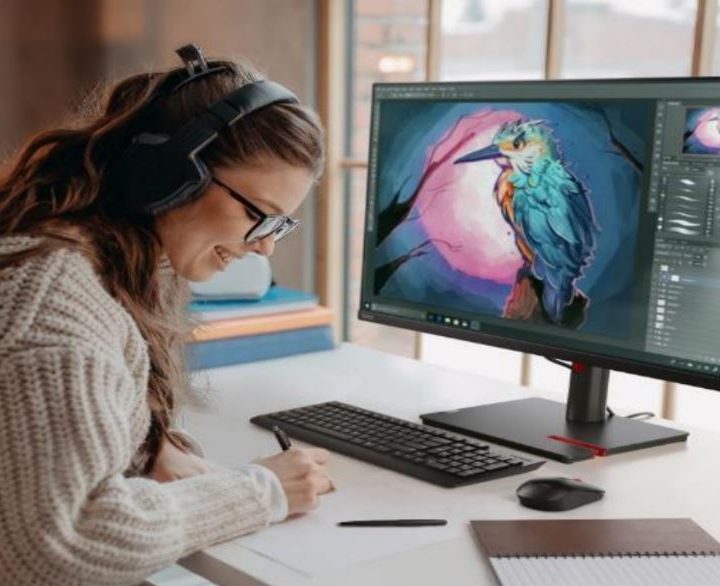 A woman smiles as she works, sitting at a desk with the Lenovo ThinkVision monitor on it.