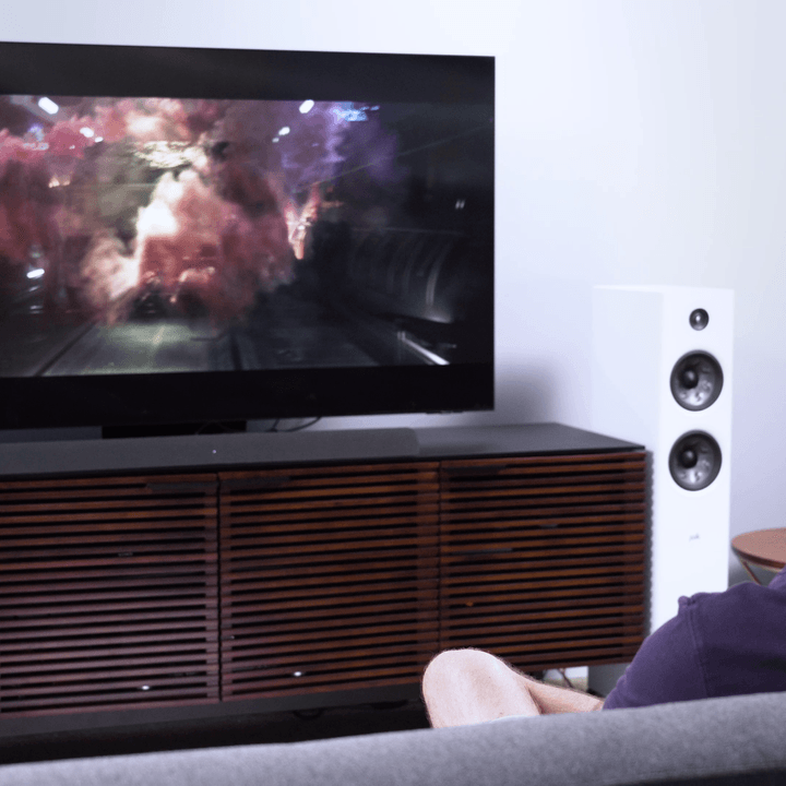 A man watching TV that is sitting on a media cabinet with the Vizio M-Series AIO soundbar under the TV.