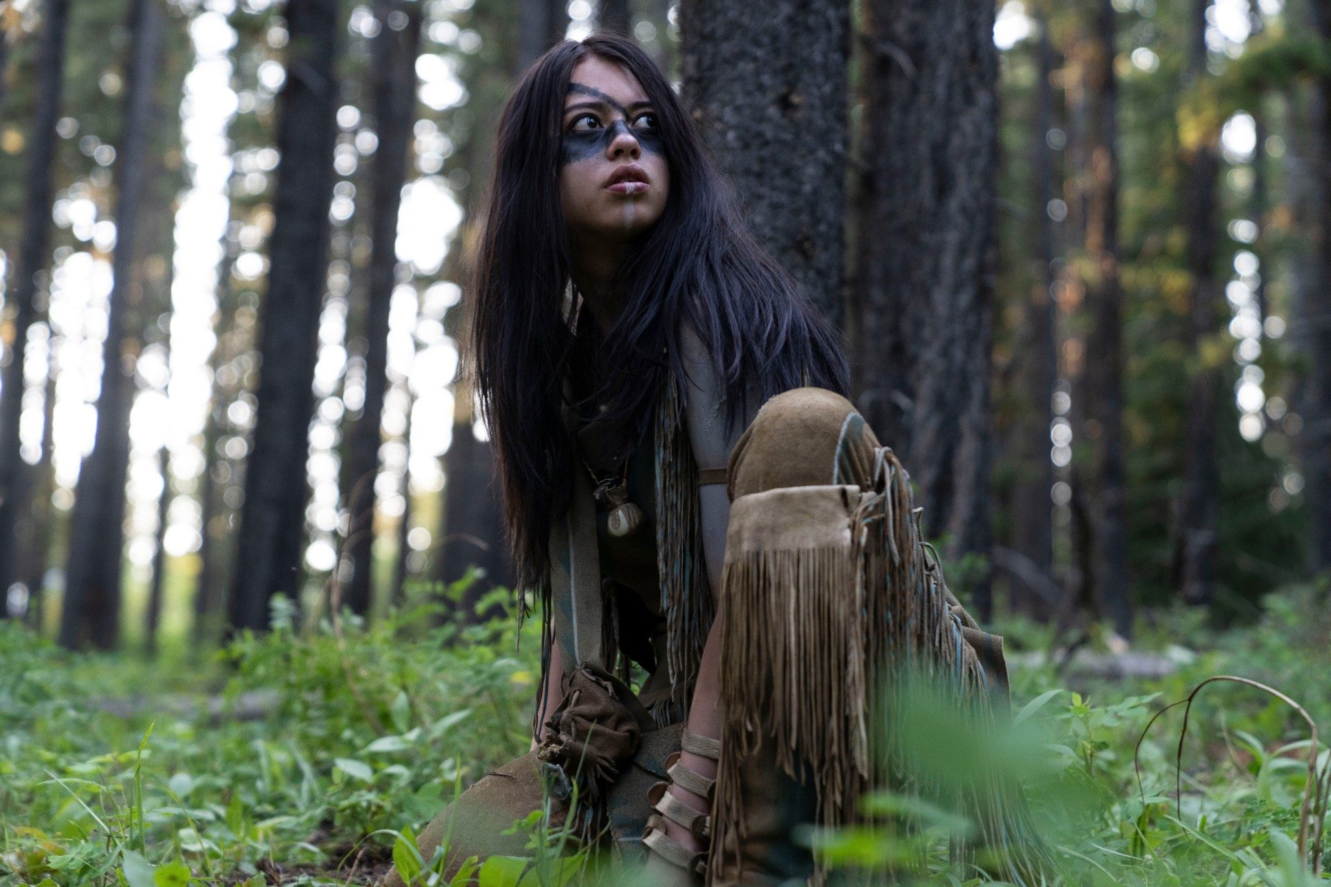 Amber Midthunder crouches in the forest, looking around, in a scene from Prey.