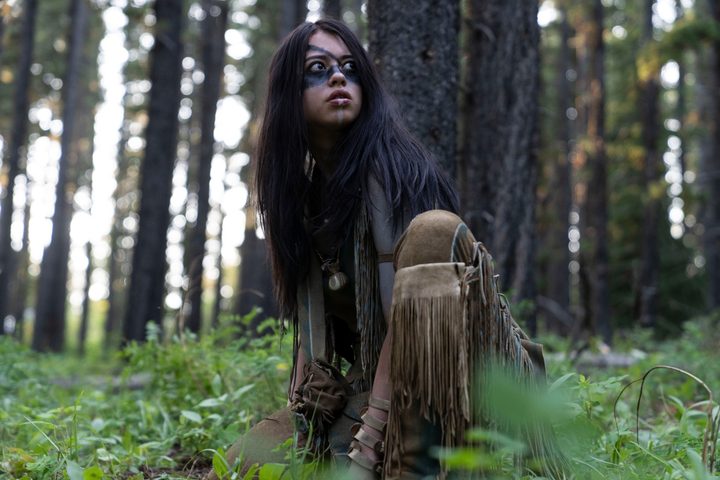 Amber Midthunder crouches in the forest, looking around, in a scene from Prey.