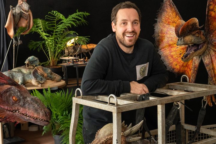 John Nolan, the live-action dinosaur supervisor on Jurassic World Dominion, standsin front of several puppets from the film.