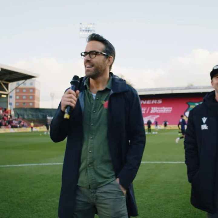 Ryan Reynolds addressing a crowd on the football field, Rob McElhenny behind him, in a scene from Welcome to Wrexham on Hulu.