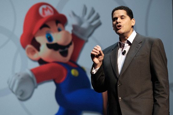 A man speakers, gesturing with his hand, in front of a large mural of Mario.