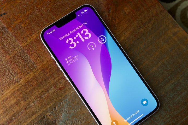 An iPhone 14 laying face-up on a wood end table. The display is on, showing the lock screen.