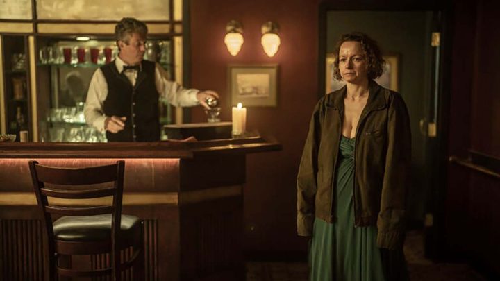 Dee wearing a dress, a bartender in the background in a scene from Tales of the Walking Dead.