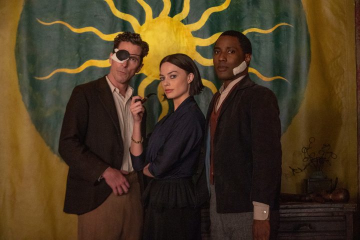 Christian Bale, Margot Robbie, and John David Washington stand in front of a sun tapestry in Amsterdam.