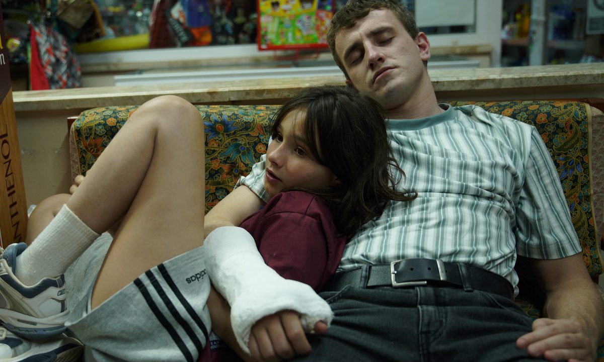 Frankie Corio sits on a couch with a sleeping Paul Mescal in A24's Aftersun.