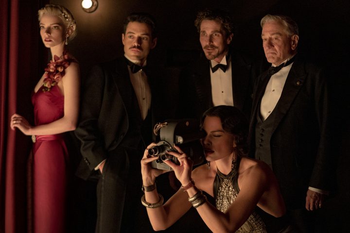 Margot Robbie holds a film camera while crouching in front of Anya Taylor-Joy, Rami Malek, Christian Bale, and Robert De Niro in Amsterdam.