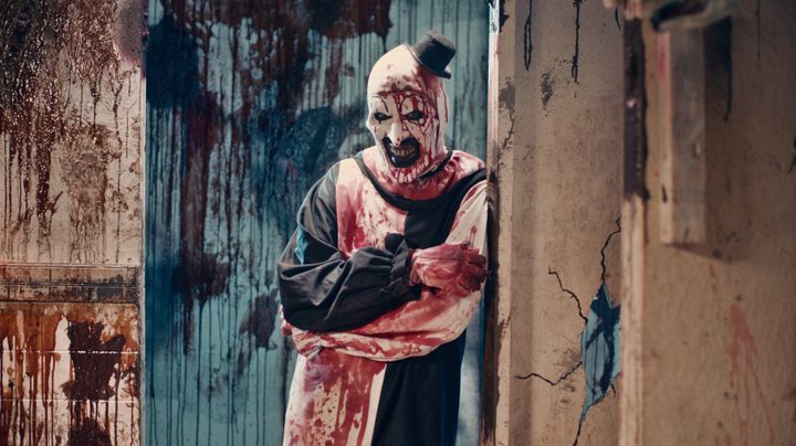 Art the Clown leans up against the wall and stares creepily in a still from Terrifier 2.