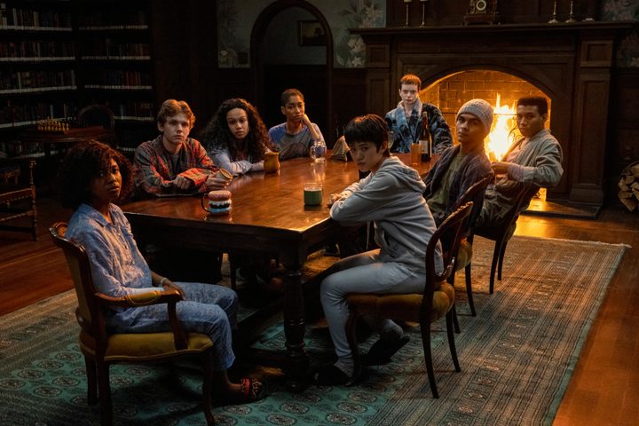 The Midnight Club kids all sit around a table together in a dimly lit library.