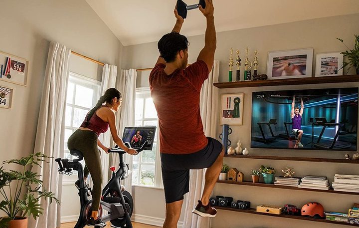 Woman riding original Peloton Bike in a family room with a man working out with weights with interactive class.