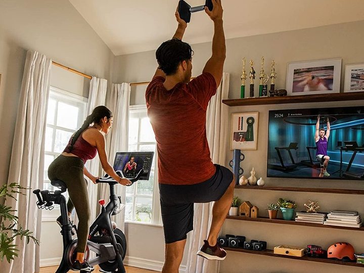Woman riding original Peloton Bike in a family room with a man working out with weights with interactive class.
