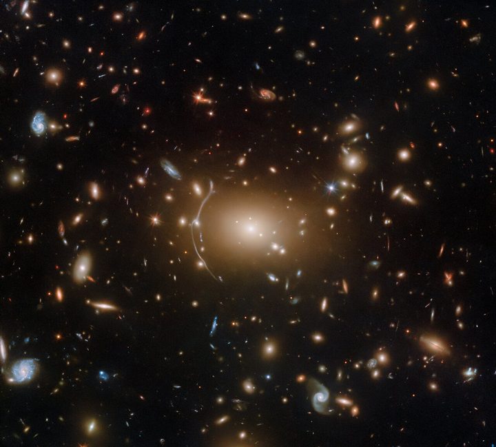Hundreds of small galaxies appear across this view. Their colours vary. Some are shades of orange, while others are white. Most appear as fuzzy ovals, but a few have distinct spiral arms. There are also many thin, long, orange arcs that curve around the centre of the image, where there is a prominent orange glow.