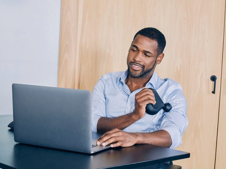 A man sitting at a desk using a Theragun mini 1.0 Handheld Percussive Massage Device on his upper arm.