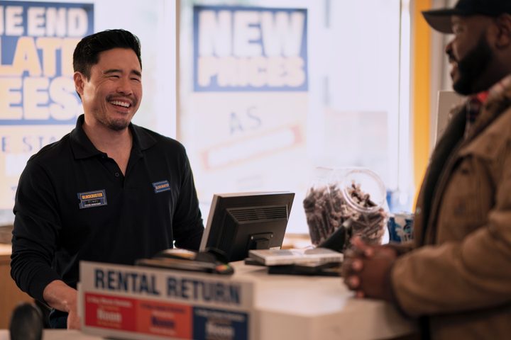 Randall Park smiles while standing behind a checkout counter in Netflix's Blockbuster.