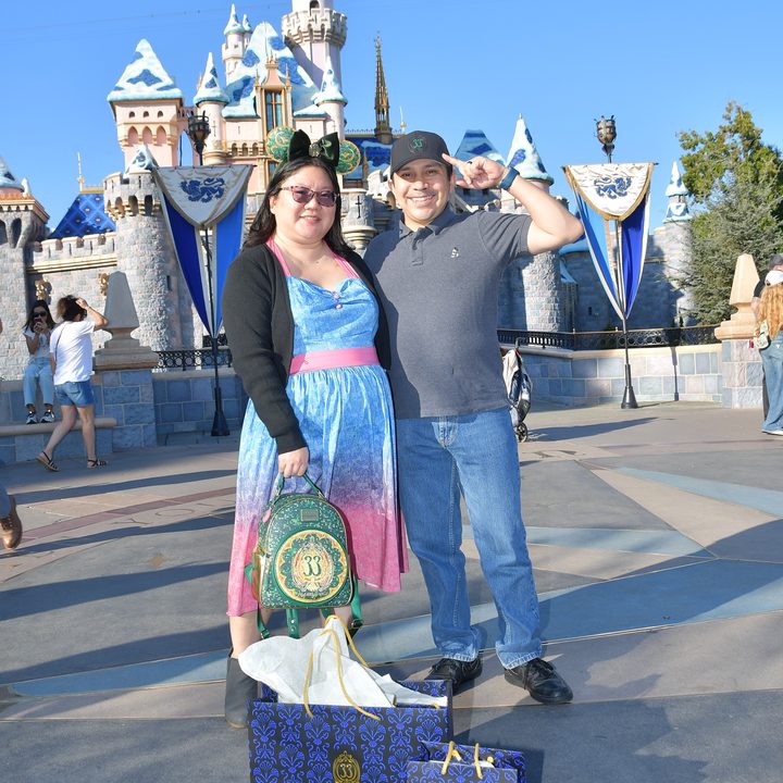Christine and husband pose in front of Disneyland castle with Club 33 shopping bags.