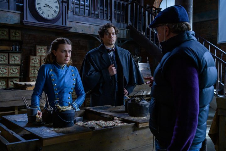 Enola Holmes 2 director Harry Bradbeer talks to stars Millie Bobby Brown and Henry Cavill on the set of the film.