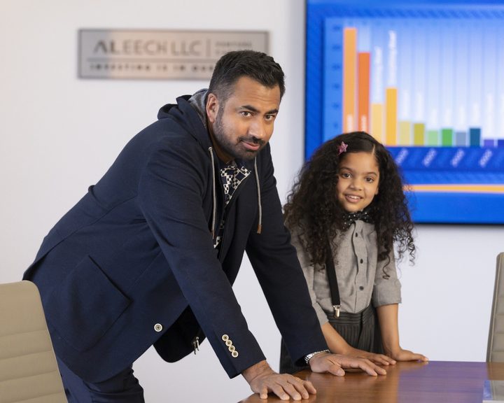 Kal Penn and Rupali Redd lean on a desk in a scene from The Santa Clauses.