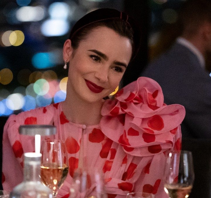Lily collins smiles while sitting at a table from Emily in Paris.