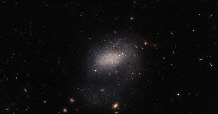 A host of astronomical objects throng this image from the NASA/ESA Hubble Space Telescope. Background galaxies ranging from stately spirals to fuzzy ellipticals are strewn across the image, and bright foreground stars much closer to home are also present, surrounded by diffraction spikes. In the centre of the image, the vague shape of the small galaxy UGC 7983 appears as a hazy cloud of light. UGC 7983 is around 30 million light-years from Earth in the constellation Virgo, and is a dwarf irregular galaxy — a type thought to be similar to the very earliest galaxies in the Universe.