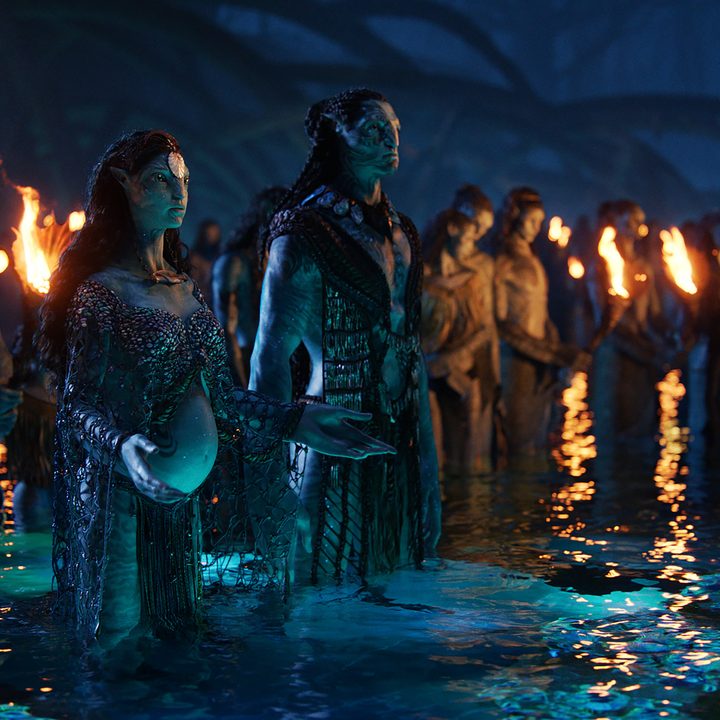 A group of Na'vi mourn in the water while flames show around them in a scene from Avatar: The Way of Water.