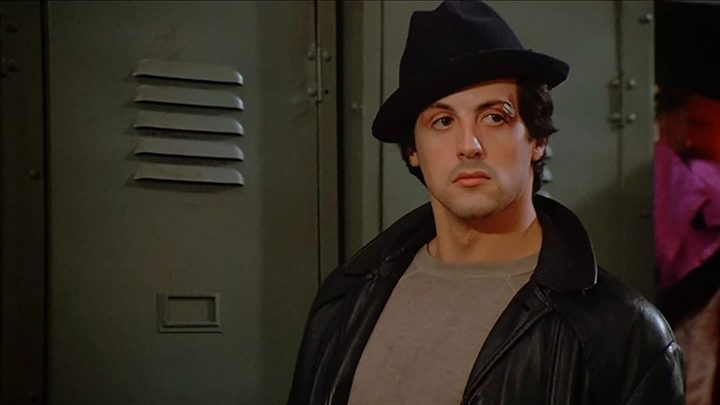 Rocky Balboa standing by a locker with a hat on, his eye bruised in a scene from Rocky.