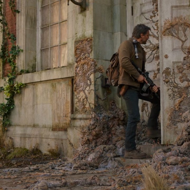 Joel holds a rifle in front of him as he enters a door covered in dried fungus in The Last of Us on HBO.
